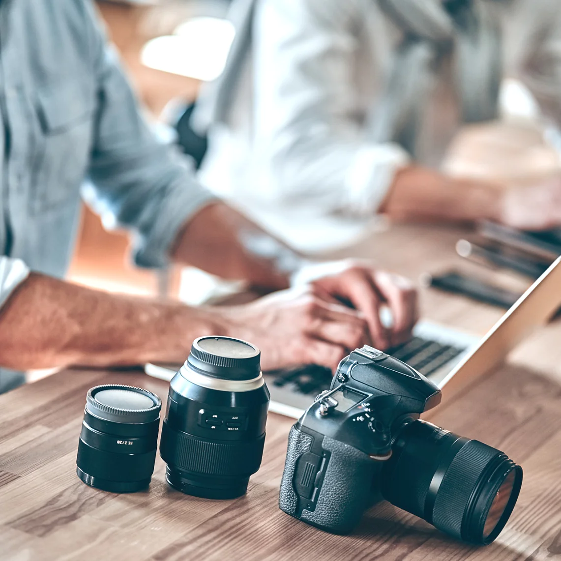 A person in a blue shirt types on a laptop at a wooden table. Next to them are three camera lenses and a DSLR camera, secured with tesa tape. Another person is partially visible working on another laptop in the background. (This text has been generated by AI)